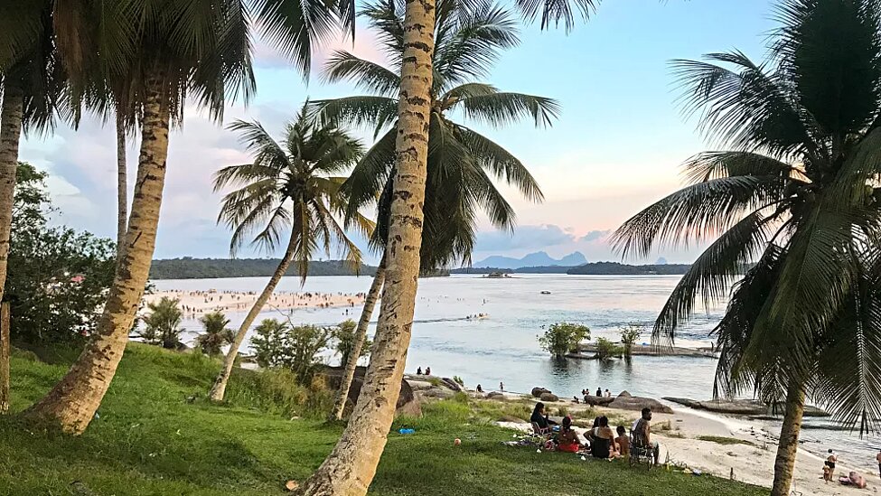 Bild på en strand med djungeln och palmer bakom på land med kanoter i vattenbrynet. Det är många invånare på stranden