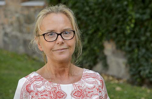 Lund University Director Susanne Kristensson is photographed outdoors. She has blond hair styled up in a bun and dark glasses. She is smiling and looking at the camera. She is wearing a short-sleeved white blouse with a red paisley pattern.